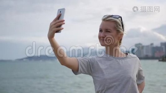 美丽的苗条女人，一个扎着辫子的金发女郎，穿着灰色的T恤，站在桥上，用手机自拍视频