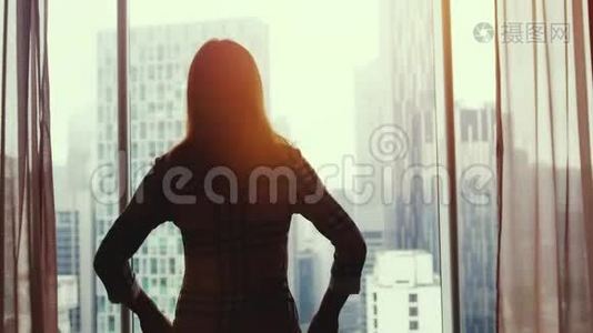 在美丽的夕阳下，年轻的黑发女子站在大窗户旁，望着城市建筑上的公寓视频