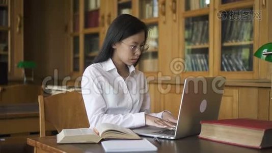 漂亮的女人正在使用笔记本电脑，看书坐在图书馆的桌子上。视频