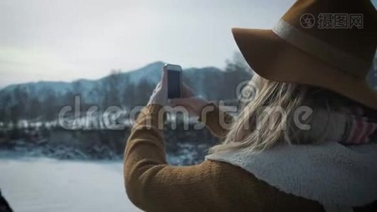 一个年轻的女孩旅行者在智能手机上拍照。 拍摄一条冰冻的冬季河流.. 一顶帽子和一个旅行者`背包视频