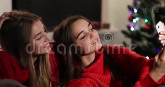 新年庆典。 4k. 两个女孩在圣诞树前坐在沙发上的智能手机上自拍视频