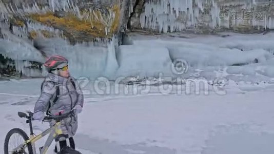 女人正走在冰窟附近的自行车旁。 有冰洞和冰柱的岩石非常美丽。 女孩是视频
