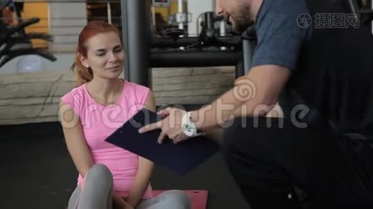 年轻的女运动员看着剪贴板，她的男教练正在健身房展示她的成绩。视频