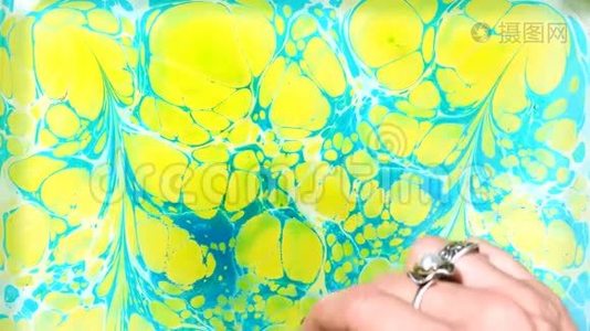 埃布鲁是在液态水面上作画的艺术。 纸大理石花纹。 女人用刷子、黄色和蓝色油漆画画。 关闭视频