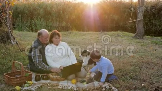 快乐的老人在日出的背景下和孙子、狗坐在格子花格子上，用电脑野餐视频