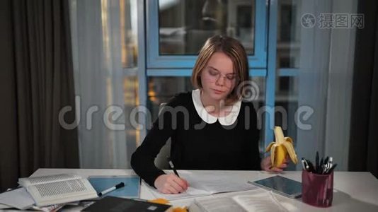 一个快乐的女学生用平板电脑吃香蕉，在笔记本上写练习。 午休时间。 她`做她视频