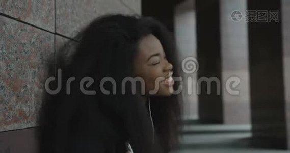 侧写。 一个情绪激动的非洲女孩正靠在大楼里，一边用耳机欣赏音乐。 4k视频