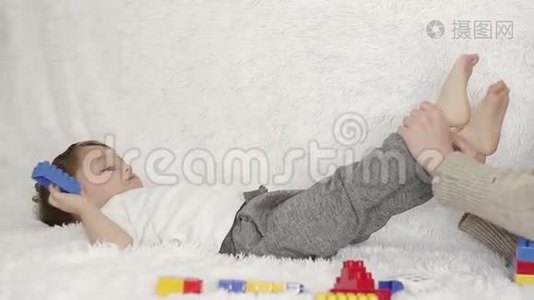 一位快乐的母亲亲吻孩子`双腿，躺在白色沙发上，背景是彩色玩具。视频
