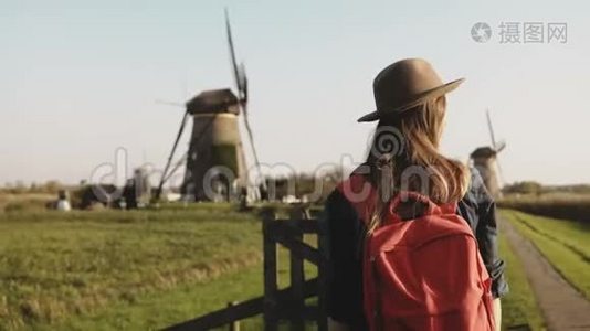 兴奋的旅游妇女在风车附近挥动手臂。 旅行女孩戴着红色背包的帽子享受乡村磨坊的风景。 4K.视频
