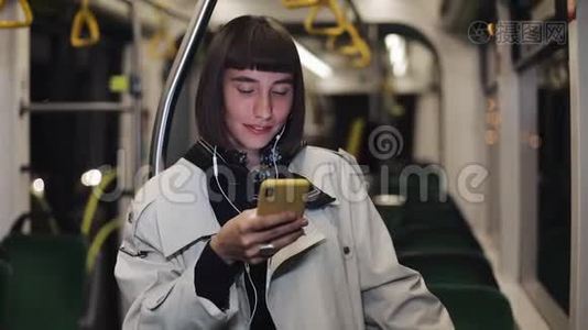 年轻时尚女性戴着耳机听音乐和在公共交通工具上浏览手机的肖像。 青年视频