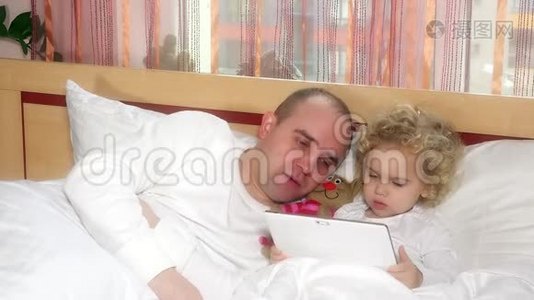 爸爸和她可爱的女儿躺在床上看着数码平板电脑视频