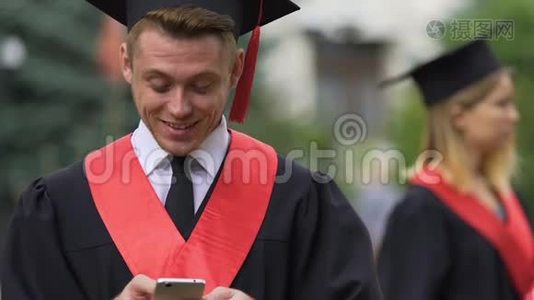 开朗的男性毕业生穿着学术服装，在智能手机上聊天，笑着视频