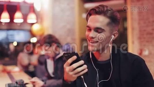 英俊的年轻人用耳机和智能手机进行视频通话。 他坐在靠近霓虹灯标志的酒吧里喝酒视频