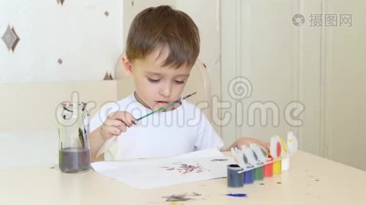 一个小男孩坐在桌子旁，用画笔在一张纸上画画。 水彩颜料。视频