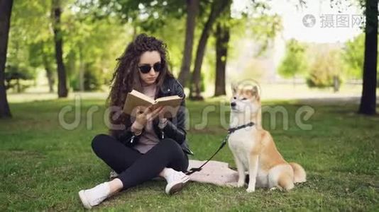 有魅力的年轻女子正坐在公园的草地上看书，而她养得很好的狗正坐在她的主人旁边。视频