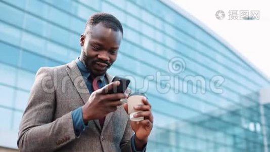 快乐的非裔美国人成功的商人在智能手机上得到了好消息。 他站在一个办公中心附近。 智能手机视频