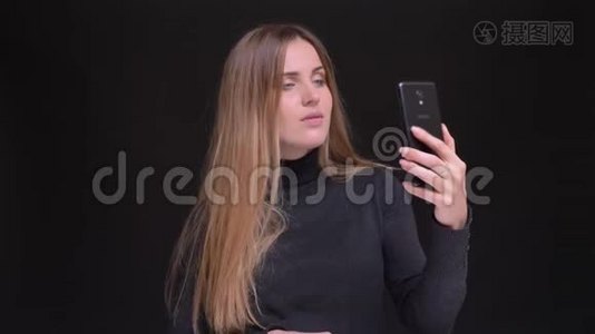 年轻的白种人长发金发金发女孩在黑色背景下使用智能手机制作自拍照片的肖像。视频