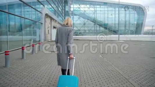 金发女孩在机场候机楼附近滚动一个手提箱-从后面看视频