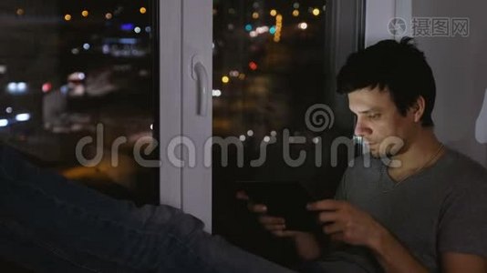 男人黑发在平板电脑上玩游戏。 在黑暗中坐在窗台上。视频