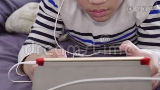 亚洲可爱的男孩在平板电脑上观看，戴着白色耳机，脸上带着微笑。视频