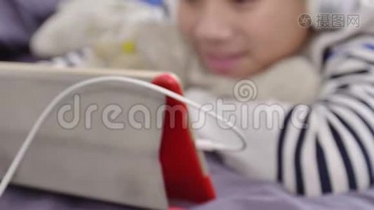 亚洲可爱的男孩在平板电脑上观看，戴着白色耳机，脸上带着微笑。视频