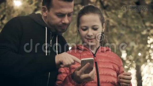 爸爸把音乐之女放在智能手机的耳机里。一个女孩在街上听一首歌跳舞视频