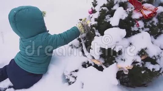 男孩，孩子躺在圣诞树附近的雪上，在户外玩装饰。视频