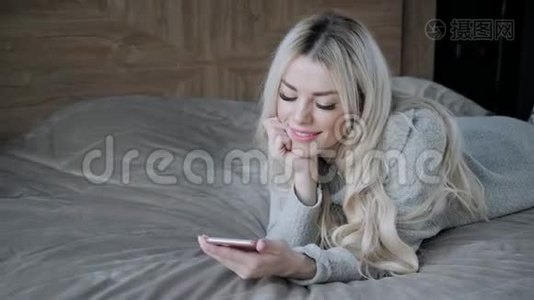 美丽的金发女人躺在床上，带着智能手机。 女孩微笑，心情好。 博客，浏览互联网视频