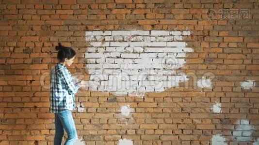 一个年轻的女孩用滚筒在她的房子里画一堵砖墙。 用滚筒粉刷墙壁。 把裸露的墙壁漆成视频