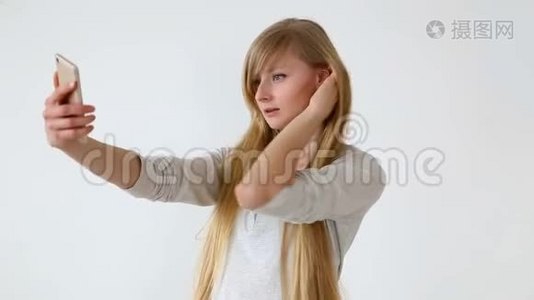 现代青年。 漂亮的长头发的欧洲女孩，金发在靠近白色墙壁的智能手机上自拍视频