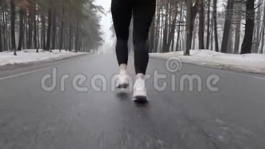 年轻迷人的白种人女孩在冬天带着耳机在雪地公园跑步。 关闭后跟镜头视频