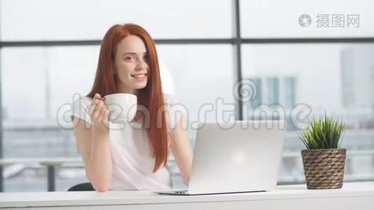 快乐的红头发女孩坐在桌子旁，手提电脑，看着相机。视频