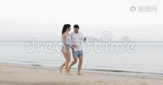 一对夫妇在海滩上用手机散步，拥抱交谈，年轻男女游客在网上交流视频