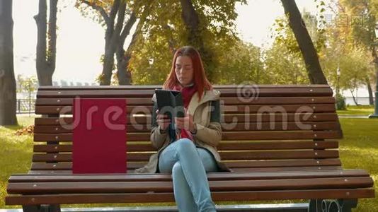 在阳光明媚的天气里，长着狐毛的女孩坐在长凳上，使用平板电脑，并在网上订购视频