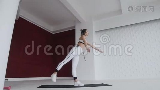 穿白色运动服的女人站在一条腿上，瑜伽练习在室内，背景宽敞的白色和红色墙壁上。 Yogi视频