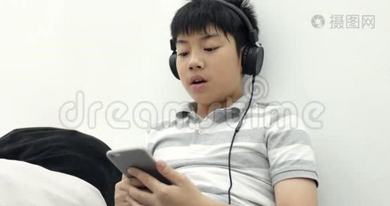 一个快乐的亚洲青少年男孩躺在清醒的智能手机和耳机和听音乐在家里的肖像。视频