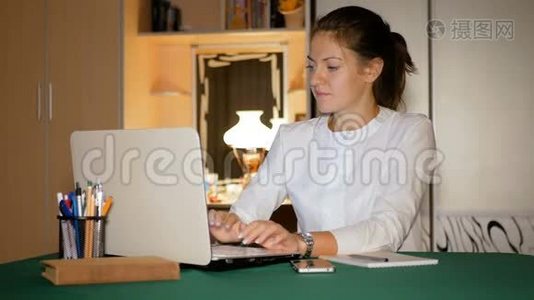 女孩自由职业者在温暖的家庭环境中为笔记本电脑工作。 布鲁内特把信息从屏幕写到视频