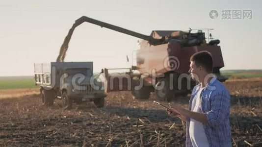 英俊的农民与平板站在联合收割机的背景。 农民使用现代技术触摸平板电脑视频