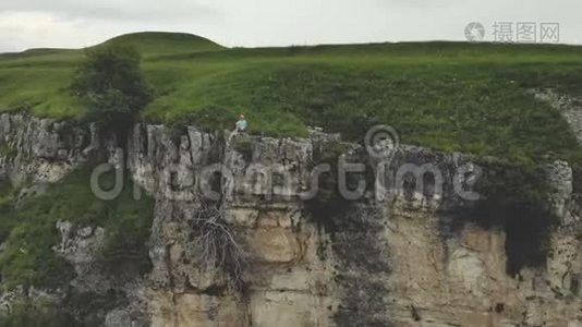 年轻的时髦女人坐在高高的岩石墙上的边缘。 空中观景。 无人机缓慢地向后飞行视频