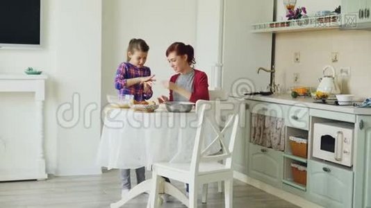 微笑的母亲和可爱的女儿一起做饼干，用面团，而坐在现代厨房在家里。 家庭视频