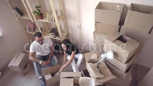 把一个年轻的家庭搬到一个新的公寓。 丈夫和妻子互相拥抱，坐在地板上，周围是盒子和视频