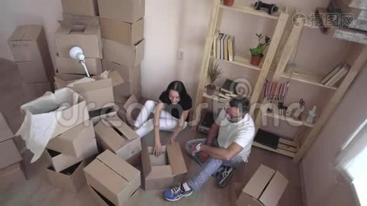 把一个年轻的家庭搬到一个新的公寓。 丈夫和妻子互相拥抱，坐在地板上，周围是盒子和视频