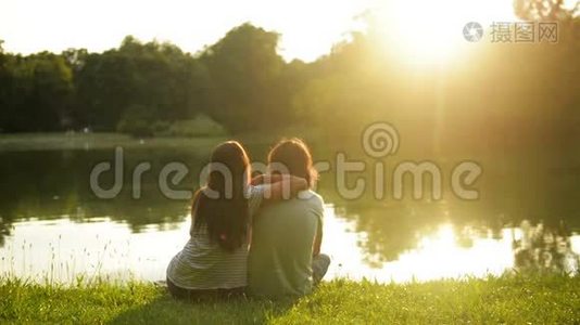 日落时，幸福的年轻夫妇坐在湖边的绿草上。 回到户外恋爱中的男女。视频