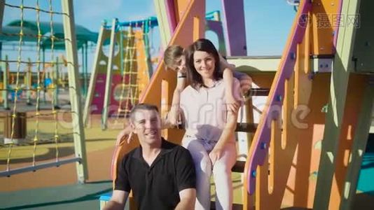 幸福的三口之家坐在孩子们`操场上滑梯。视频
