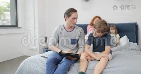 父亲和儿子用数码平板电脑坐在母亲的床上，女儿抱着笔记本电脑家庭在一起视频