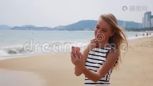 年轻漂亮的苗条女人，一头金发，穿着黑白连衣裙，在海边的手机上自拍视频