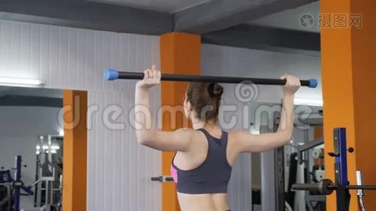 年轻漂亮的运动女孩在健身房举起杠铃棒，肩部锻炼，背面观看。 60英尺视频