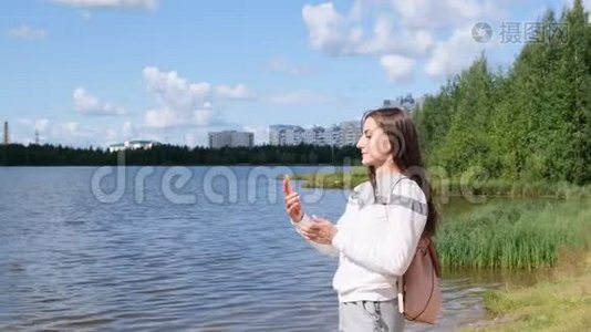 带背包的漂亮女孩在湖边自拍，并在Skype上用手机交谈。视频