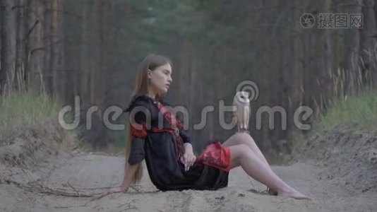 一个穿着黑色连衣裙的长发女孩光着脚坐在一条奉承的小路上，膝盖上坐着一只浅棕色的猫头鹰，慢吞吞的视频