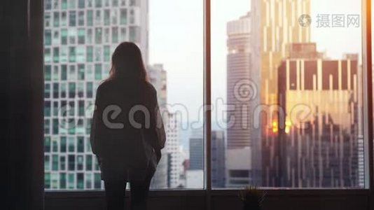 夕阳西下，年轻忧郁的女人站在大窗户旁，望着她在城市建筑上的公寓。 慢动作视频